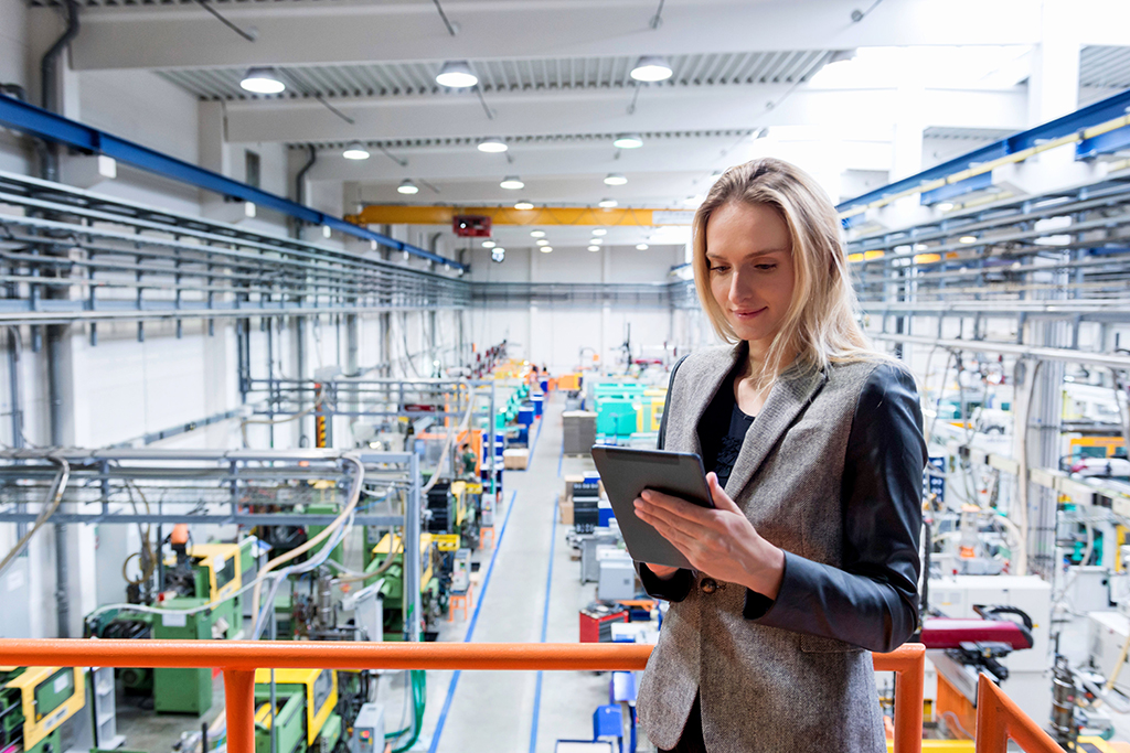 Horizontal color image of blond business female worker standing on balcony on top of large factory, holding digital tablet and examining the production online. Focus on attractive businesswoman, futuristic machines in background.